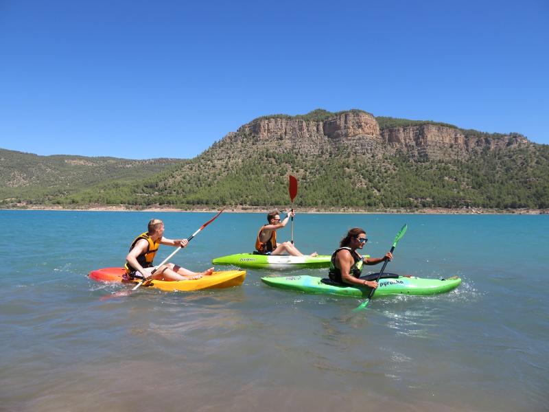 Três pessoas praticam canoagem em águas calmas com paisagem montanhosa ao fundo como parte de atividades de lazer num curso de espanhol.