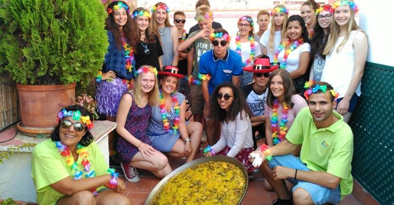 Grupo de pessoas vestido com acessórios festivos posa junto a uma grande paella como parte de uma celebração em um curso de espanhol.