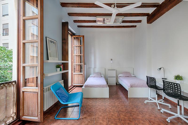 Quarto luminoso com duas camas individuais, mesas de estudo e janelas amplas, acomodando estudantes de um curso de espanhol.