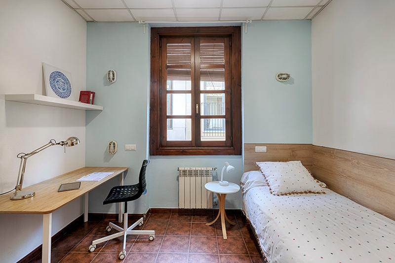 Quarto individual com cama, mesa de estudo e decoração simples, ideal para estudantes de um curso de espanhol.