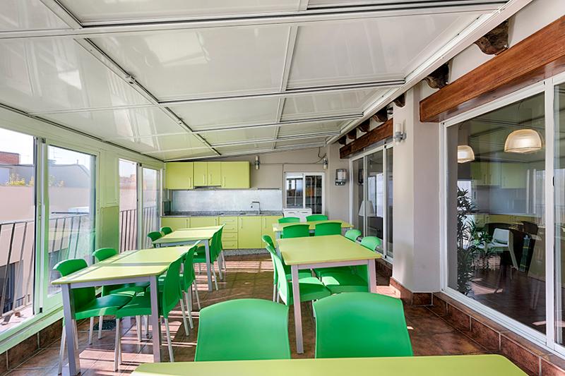 Sala de refeições iluminada com mesas e cadeiras verdes e uma área de cozinha, destinada a estudantes de um curso de espanhol.
