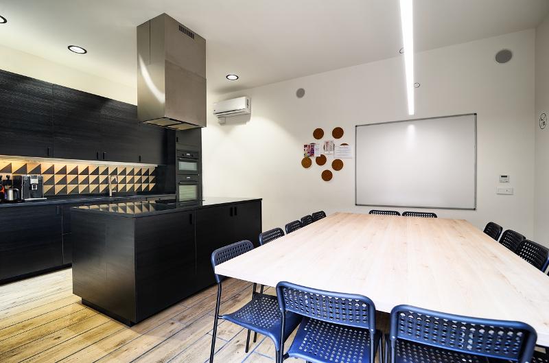 Sala moderna com cozinha equipada e mesa ampla com cadeiras, ideal para atividades em grupo num curso de espanhol.