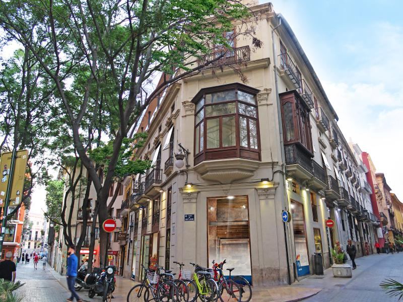 Edifício histórico numa esquina com janelas grandes e varandas em madeira, localizado numa rua movimentada com bicicletas estacionadas, onde se realiza um curso de espanhol.