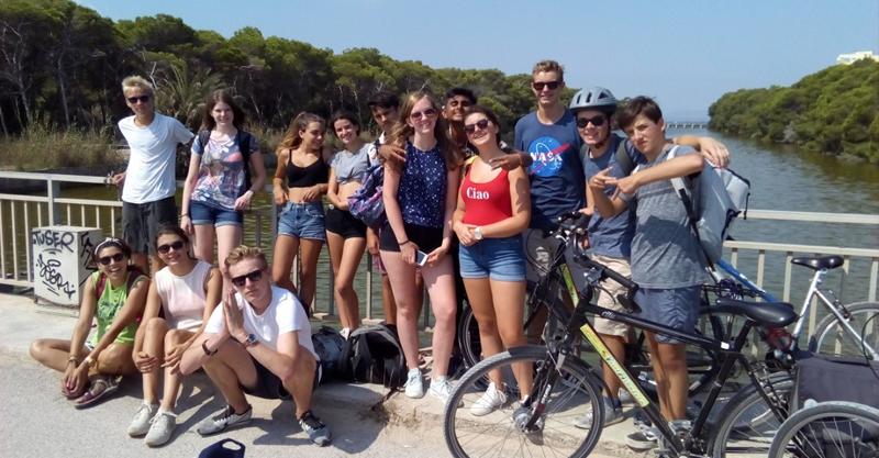 Grupo de jovens posa junto a uma ponte com bicicletas durante uma atividade ao ar livre de um curso de espanhol.