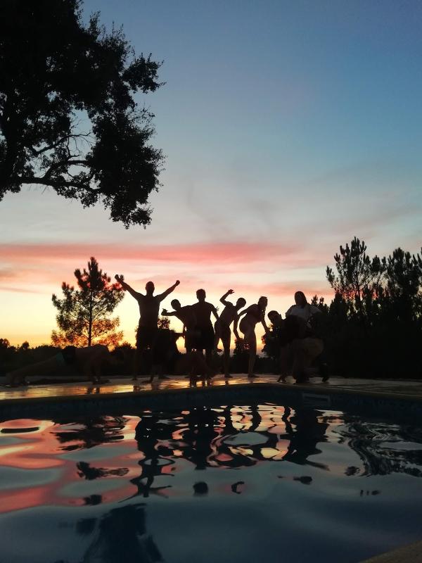 Um grupo de jovens está junto à beira de uma piscina, com o pôr do sol ao fundo, criando silhuetas vibrantes enquanto posam e celebram no ambiente do campo de férias setembro. A água reflete o céu pintado em tons de rosa e laranja, dando um toque mágico ao momento..