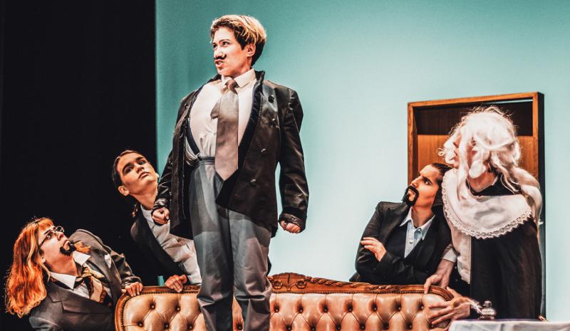  Jovens participam numa encenação teatral durante o campo de férias setembro. Vestidos de forma excêntrica e com bigodes postiços, interpretam personagens com entusiasmo e expressão intensa, destacando-se um deles em pé no sofá, assumindo uma postura teatral e confiante.