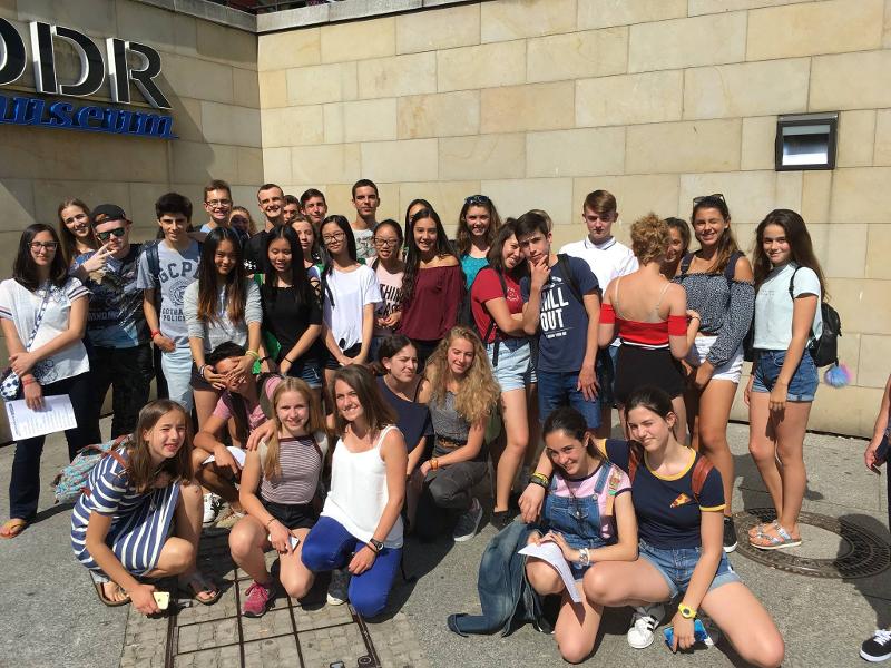Grupo de estudantes a sorrir em frente ao logo da escola de línguas, celebrando a experiência do seu curso de inglês na alemanha.