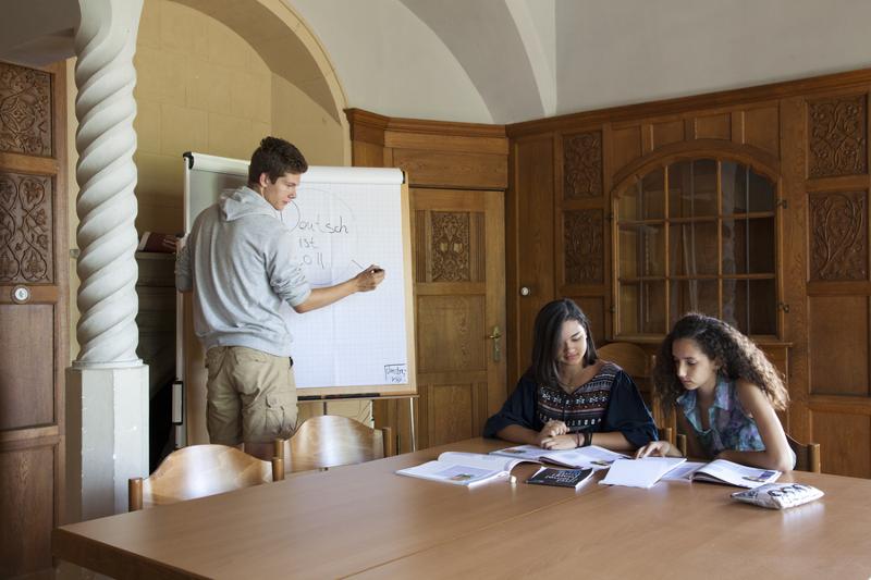 Três alunos, duas raparigas a estudar e um rapaz a escrever no quadro, envolvidos na aprendizagem durante o seu curso de inglês na alemanha.