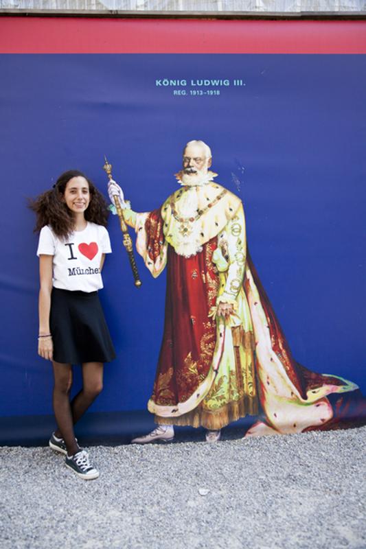 Rapariga com blusa "I love Munich" ao lado de uma imagem de uma figura histórica, sorrindo enquanto desfruta do seu curso de inglês na alemanha.