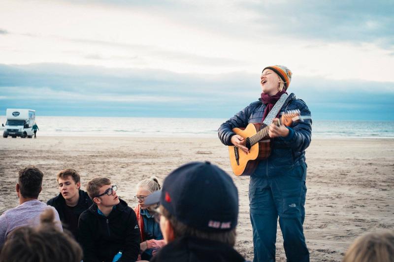 Jovem na praia a tocar guitarra, desfrutando da música e do ambiente enquanto faz o seu curso de ingles na irlanda.