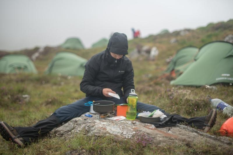 Menino a cozinhar no chão da montanha, ao lado da tenda, aproveitando o ambiente natural enquanto faz o seu curso de ingles na irlanda.