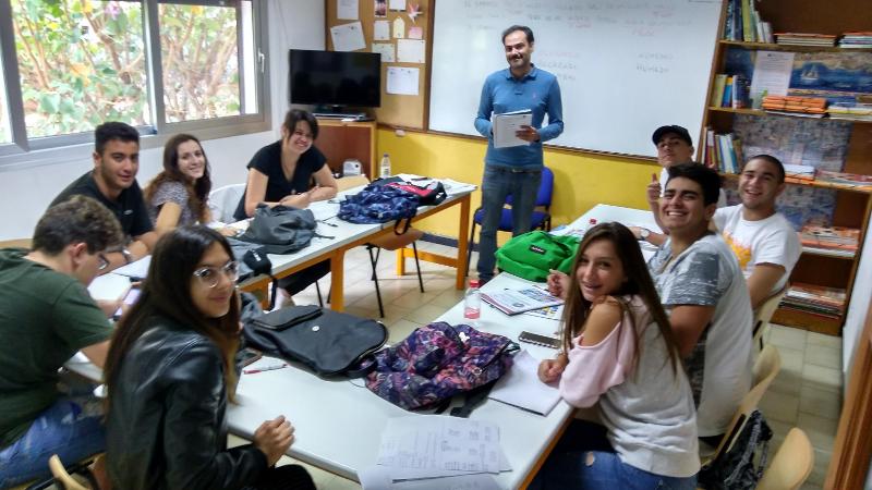 Sala de aula com grupo de estudantes sorridentes e professor, todos envolvidos num curso de lingua espanhola.
