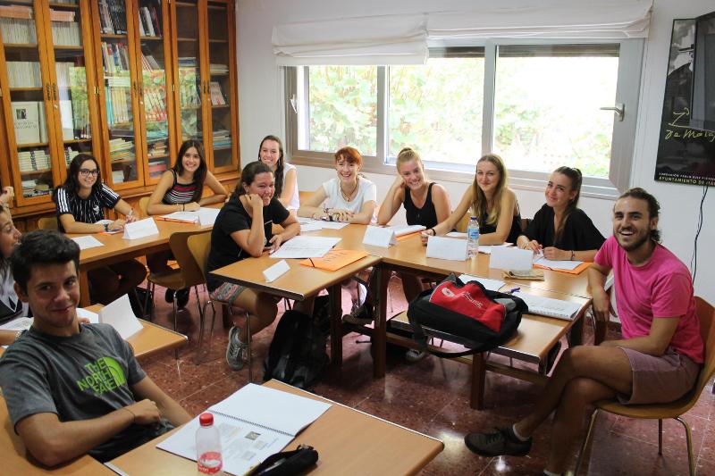 Grupo de estudantes sorridentes sentados em círculo numa sala de aula, participando num curso de lingua espanhola.