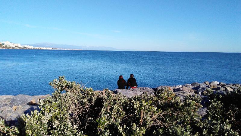 Duas pessoas sentadas numa costa rochosa com vista para o mar azul profundo, aproveitando momentos tranquilos durante um curso de lingua espanhola.
