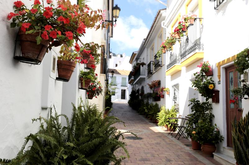Rua estreita com casas brancas e vasos floridos em varandas, proporcionando um ambiente encantador para um curso de lingua espanhola.