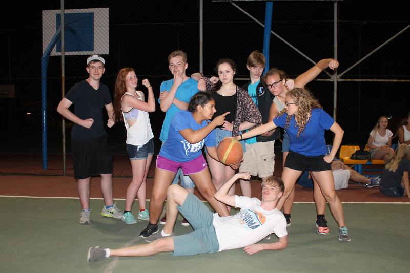 Grupo de jovens a posar de forma divertida num campo de basquetebol à noite, capturando momentos de diversão durante um curso de espanhol.