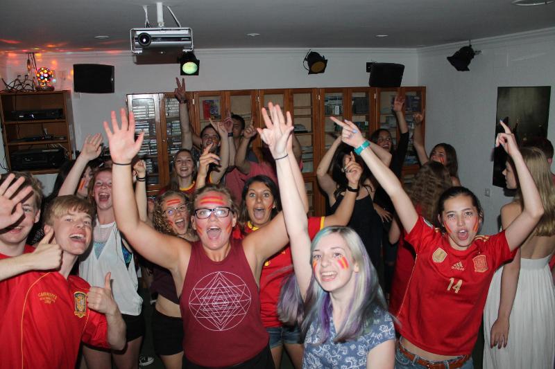 Grupo de estudantes a dançar e celebrar num ambiente festivo com camisolas de futebol, durante um evento social de um curso de espanhol.