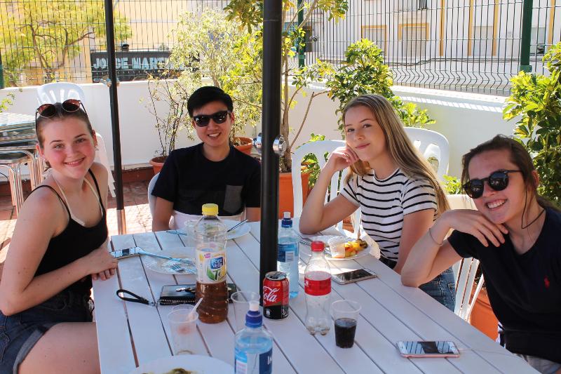 Grupo de estudantes sorridentes a desfrutar de uma refeição ao ar livre, num ambiente relaxante durante o seu curso de espanhol.