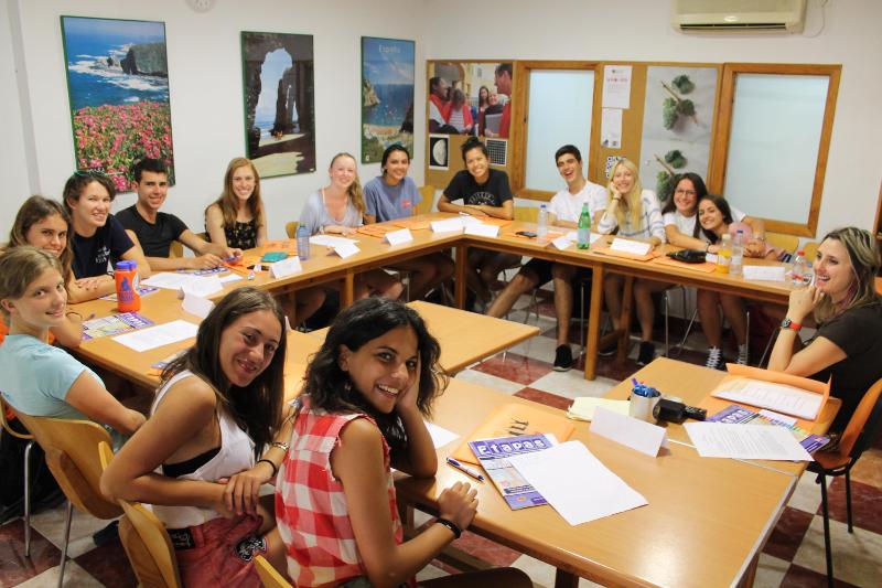 Alunos sorridentes participando de uma aula de espanhol em grupo, com material de estudo espalhado pelas mesas nos cursos de linguas em espanha.