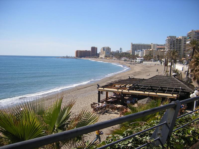 Vista da praia com espreguiçadeiras e o mar calmo, perto de edifícios, ideal para relaxar após os cursos de linguas em espanha.