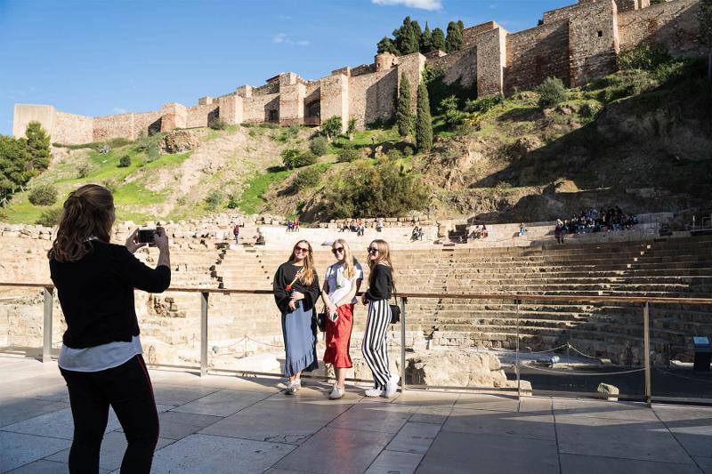 Um grupo de quatro jovens mulheres posa alegremente numa antiga arena com um castelo histórico ao fundo, enquanto uma delas tira uma fotografia, ideal para um curso de línguas no estrangeiro.